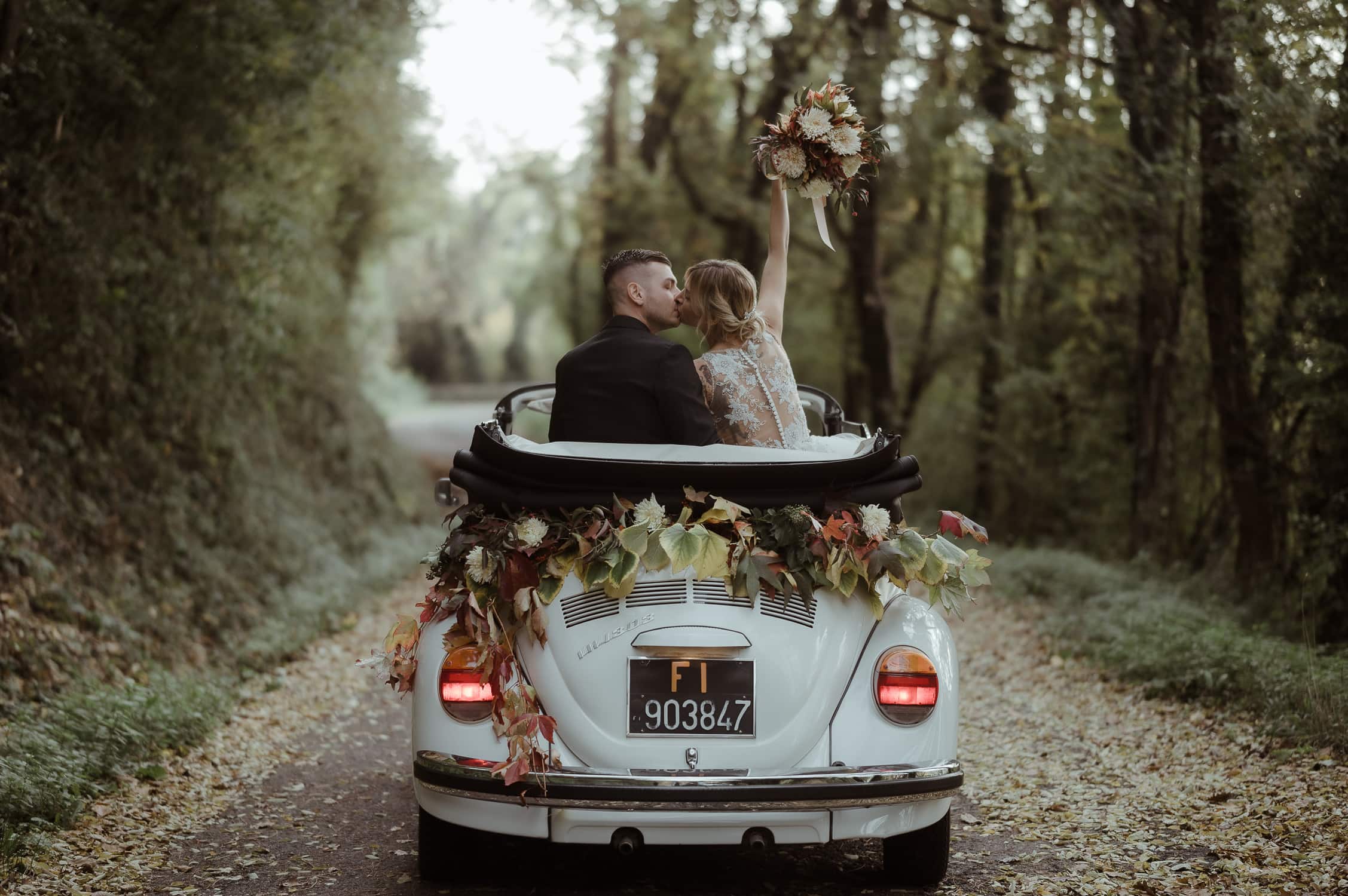 Bride and groom on a vintage VW white car with flowers while kissing in a forest in Italy | Elopement Definition